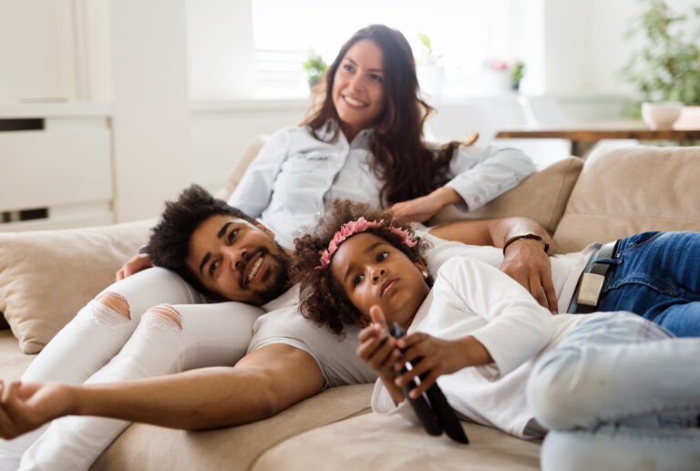 Mother, father and daughter lounge on a couch watching TV in a brightly lit living room.